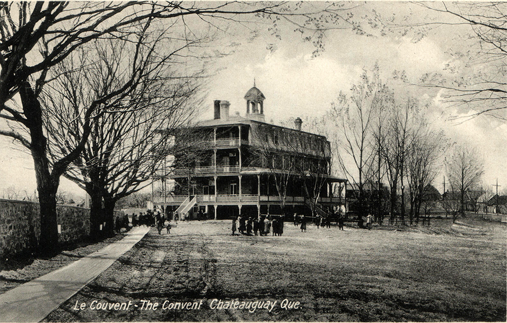 Black and white photo of the convent of the Congregation of Notre Dame. Convent girls are in front of the low wall separating the convent from the parish enclosure, on the left.