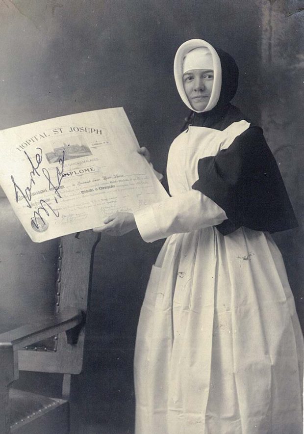 Black and white picture of Sister Marie-Louise LePailleur, in the habit of a Sisters of Providence with a nurse's apron holding her nursing diploma.