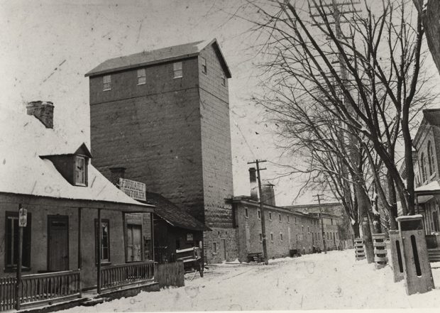 Black and white picture of a silo several stories high and a long stone building with a tall chimney as part of the Dawes Brewery facility alongside residential homes.