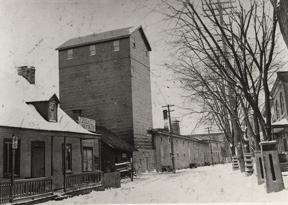 Black and white picture of a silo several stories high and a long stone building with a tall chimney as part of the Dawes Brewery facility alongside residential homes.