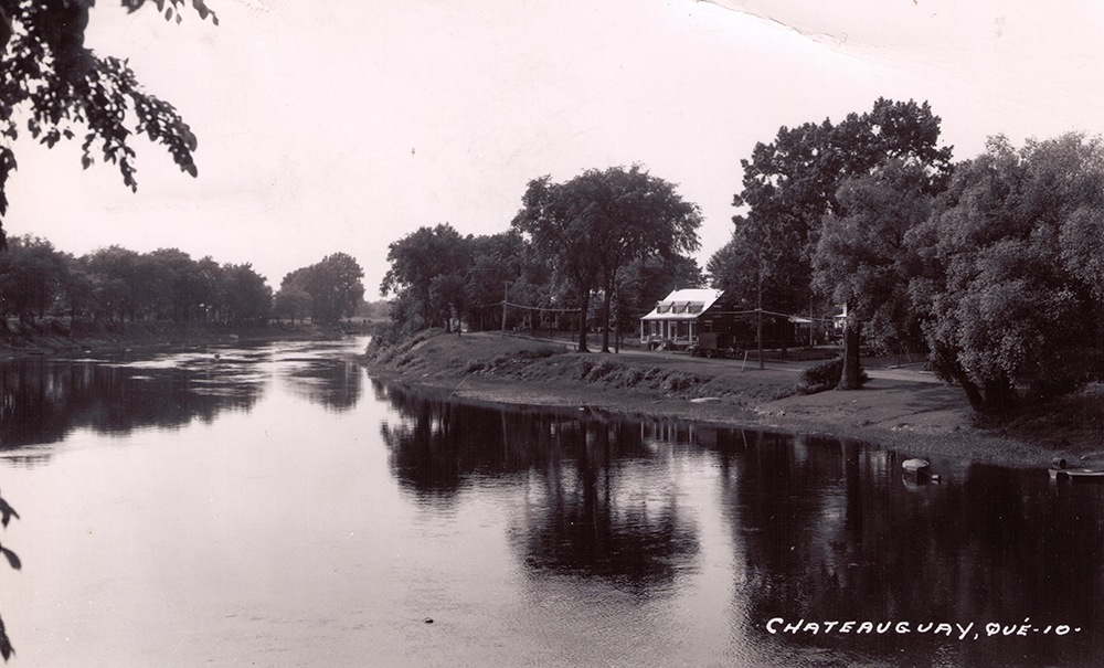 Black and white picture of the Maison Gravel. The Chateauguay River is in the foreground and several trees line the banks.