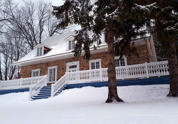 Color picture of the Maison LePailleur taken in winter. The balcony is blue with a white railing. There are two large conifers in the foreground.