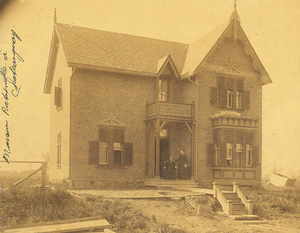 Sepia picture of Philomène Dalton on the stoop of the family house with her son, the priest Georges-Marie LePailleur. There is a part of a fence and a small staircase in the foreground but the rest of the landscape is rather empty.