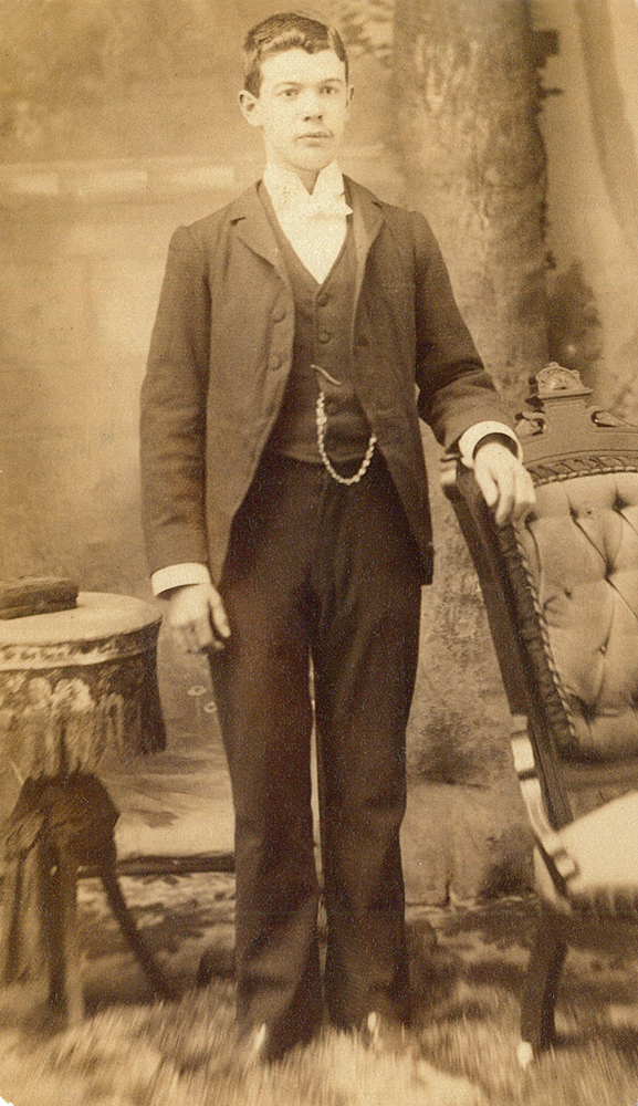 Sepia picture of young Armand LePailleur posing in a photo studio. He is standing on a fur rug. He is wearing a suit and a pocket watch.