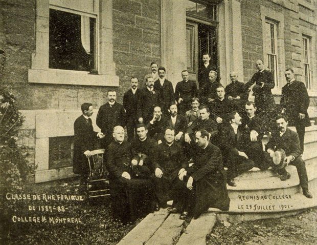 Black and white picture of a group of about 20 men in front of the Collège de Montréal. Some of them wear a cassock, others wear a suit. Those in the first row are seated, the others are standing.