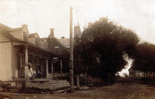 Black and white picture of Principale Street in Chateauguay. Houses are on the right side of the dirt road. A woman in a long dress is on the front porch.