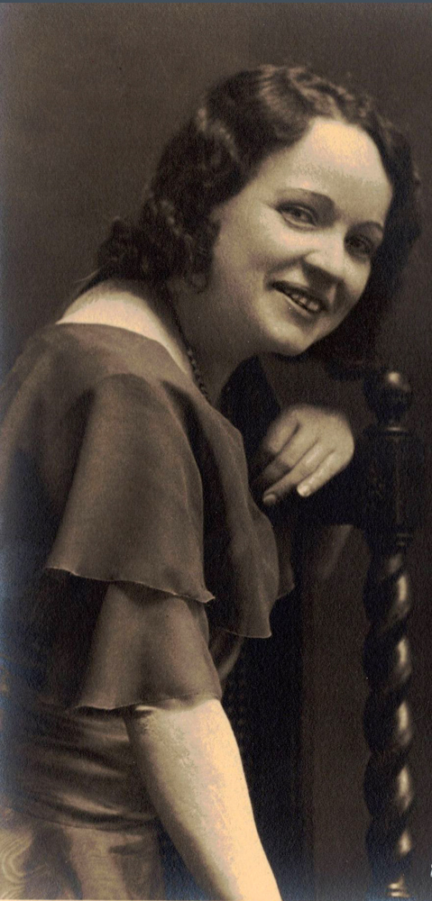 Black and white picture of Marie-Rose Descarries sitting sideways on a wooden chair. Her left arm is resting on the backrest.