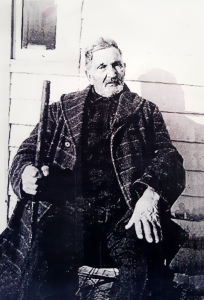 Older man seated in front of building while holding a stick