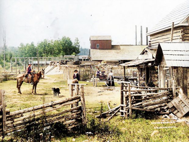 Man on horseback, dog and buggy in front of wooden buildings and fences