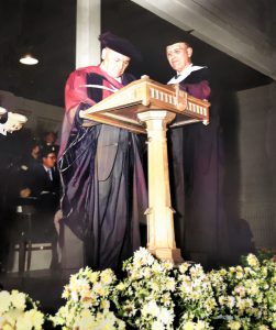 Two men in academic gown and cap standing at podium with flowers in foreground