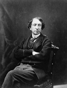 Black and white portrait of Sir John A. Macdonald, a middle-aged man with dark hair, posing in a chair in a studio, his arms folded across his chest.