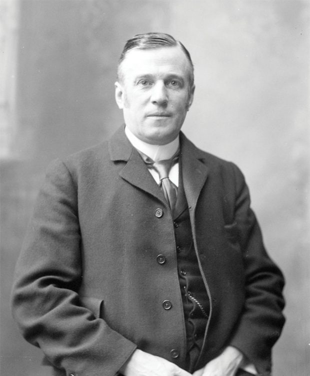 A black and white portrait of Sir Joseph Pope, from head to hips, his arms folded in front of him, wearing a three-piece suit and tie, a watch chain and high collar. He is looking directly at the camera.