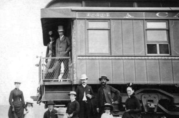 A black and white photograph of a couple (Sir John A. and Lady Agnes Macdonald) standing on the platform of a train car, while a small crowd of eight people stand below them.