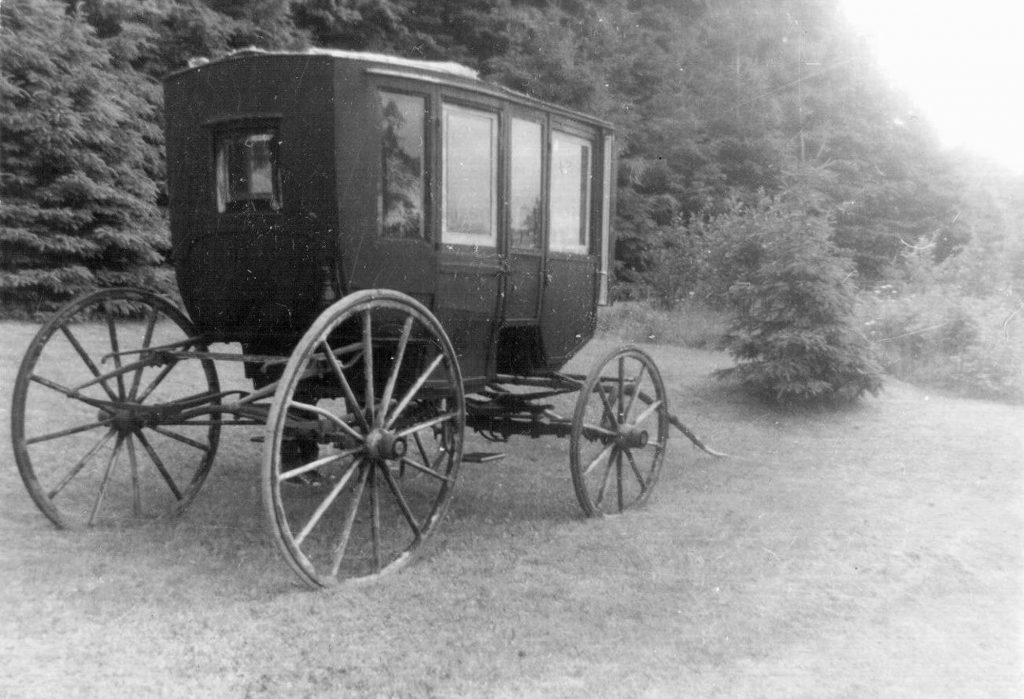 A black and white photograph of a one-horse carriage, with a covered roof and a door and windows on it, and four large wooden wheels. It is resting on a lawn with trees behind it.