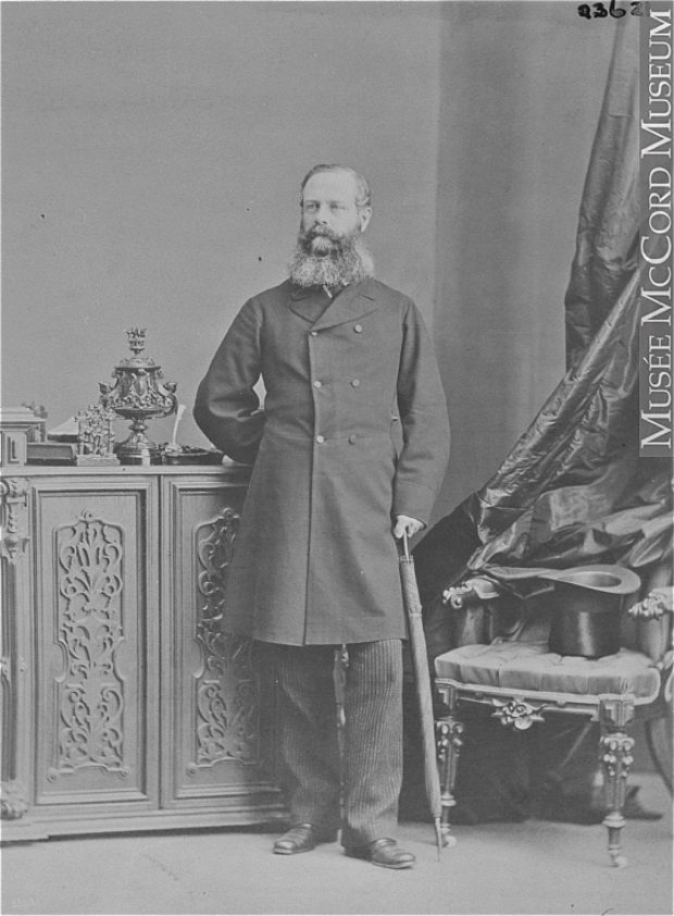 Black and white photograph of a man (Lord Monck) standing, leaning on a folded-up umbrella, his other hand held behind his back. There is a chair next to him on which sits a top hat. The man has a full beard and is dressed in a double-breasted woolen overcoat.