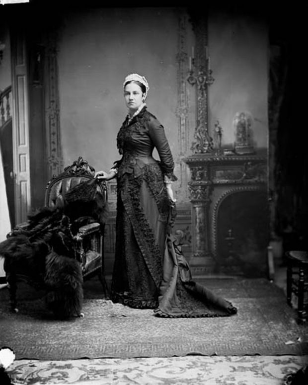 Lady Agnes Macdonald dressed in a long black dress standing in front of a hearth, next to a fur coat which is draped over a chair.