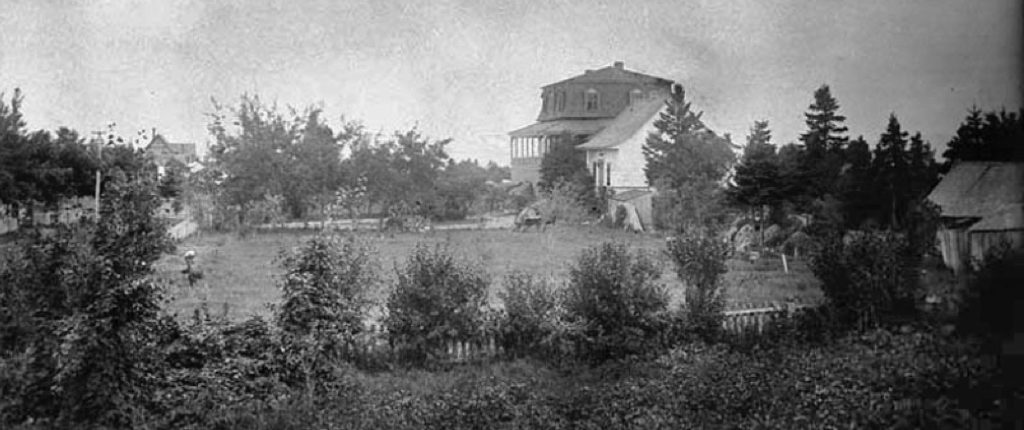 Black and white photo of summer home Villa Les Rochers, c. 1883, and partial grounds or front lawn, edged by evergreen trees. A wooden shed is in the right foreground and in the distance there is a peaked roof of the neighbouring cottage.