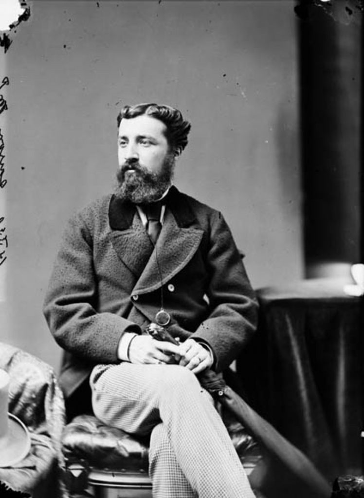 A black and white photograph of Sir Adolph Caron, a young bearded man seated, holding a closed umbrella next to his crossed legs, looking to the left of the viewer. He wears a double-breasted wool coat and a dark tie.