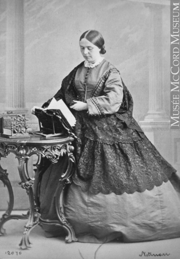 Black and white photo of a woman (Lady Frances Monck) standing, a wide full-length skirt draped with lace; she is turning the pages of a book which is open on a stand in front of her.