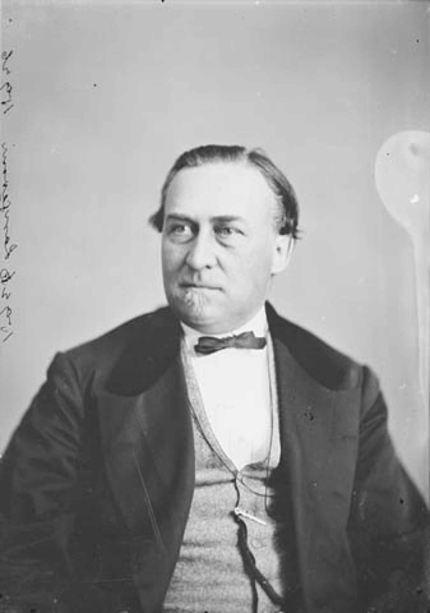 Black and white portrait of Sir Hector Langevin, a middle-aged man, clean shaven but for a small beard on his chin, dark hair, wearing a jacket and vest and bow tie, looking to the viewers left as though he's focused on something.