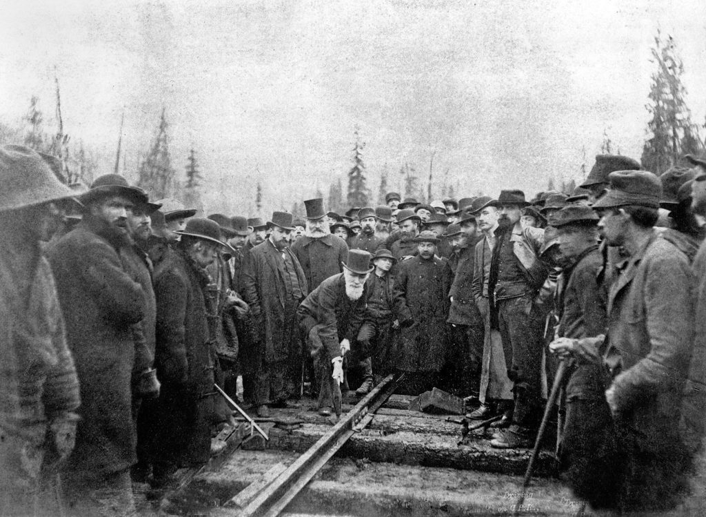 A black and white photo taken in 1885, with over a hundred gentlemen in dark woolen suits surrounding one, Sir Donald A. Smith, who is poised above a piece of railroad track, wearing a top hat and holding a large hammer against the track, about to drive the last railroad spike for the Canadian Pacific Railway.