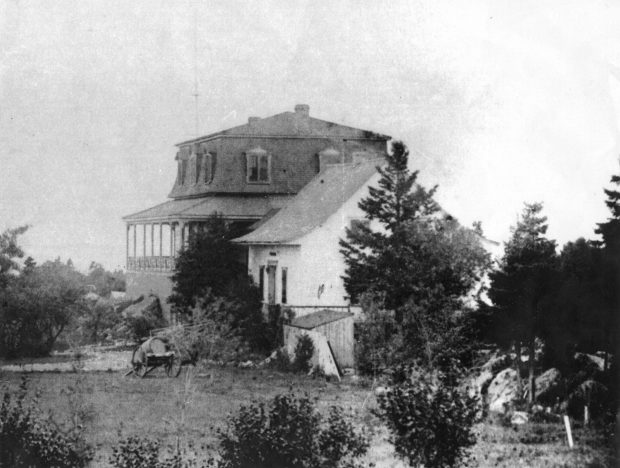 Black and white photo of Villa Les Rochers, a house that looks to be in two parts: a single plain A-frame farmhouse, with a large addition featuring a mansard roof on the far side. Trees and shrubs surround the house.