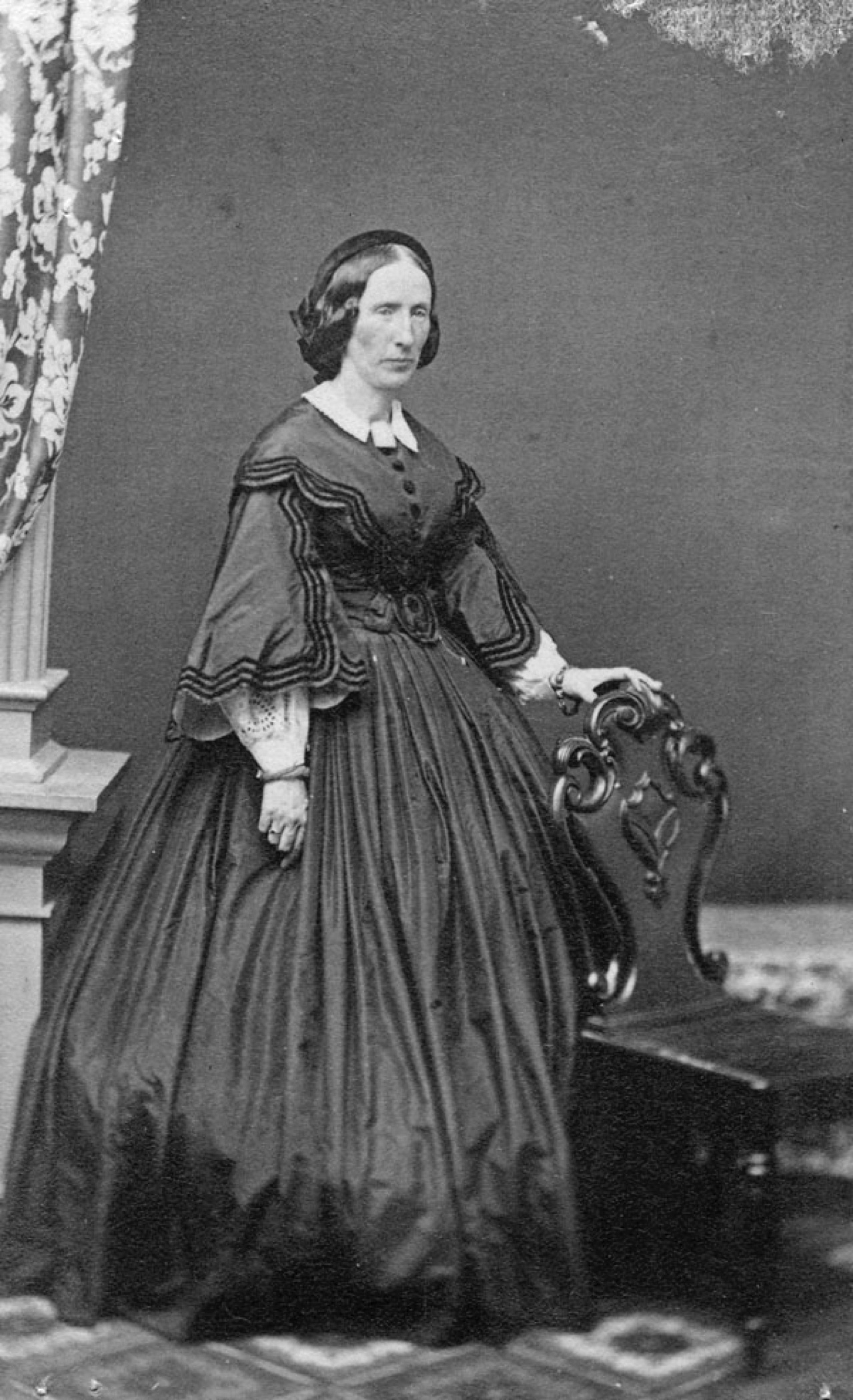 A formal black and white portrait of Louisa Macdonald, a woman standing in a full-length black dress, holding one hand on the back of a chair. Her dark hair is parted in the middle and arranged at the back. Her expression is distant and serious.