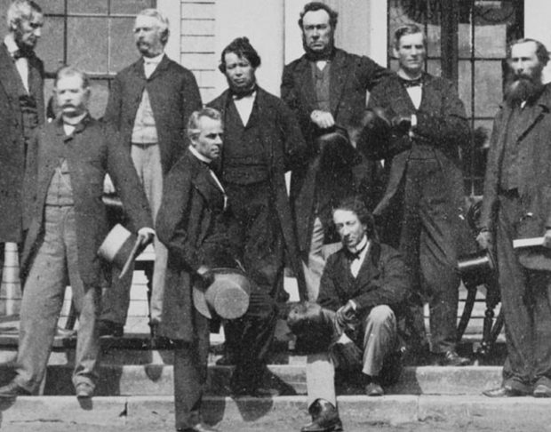 Sir John A. Macdonald sits casually on the steps in front of Charlottetown's Province House, where his Fathers of Confederation all stand around him.