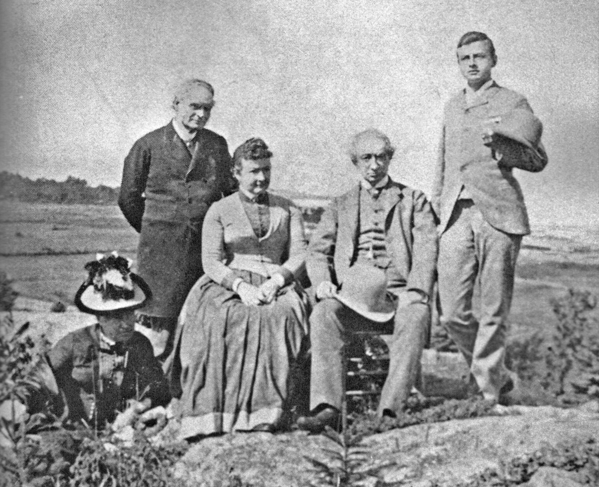 A grainy, black and white photo of five adults (Sir John A. Macdonald, Lady Agnes Macdonald and members of the Tilley family) standing and seated on some rocky ground, with the Saint Lawrence River behind them.
