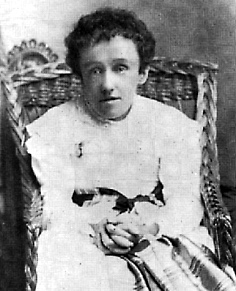 Black and white photograph of Mary Macdonald, a thin young lady leaning forward in a wicker chair, with her hands clasped on her lap. She wears a white dress with a black sash; her hair is dark and her expression vacant.