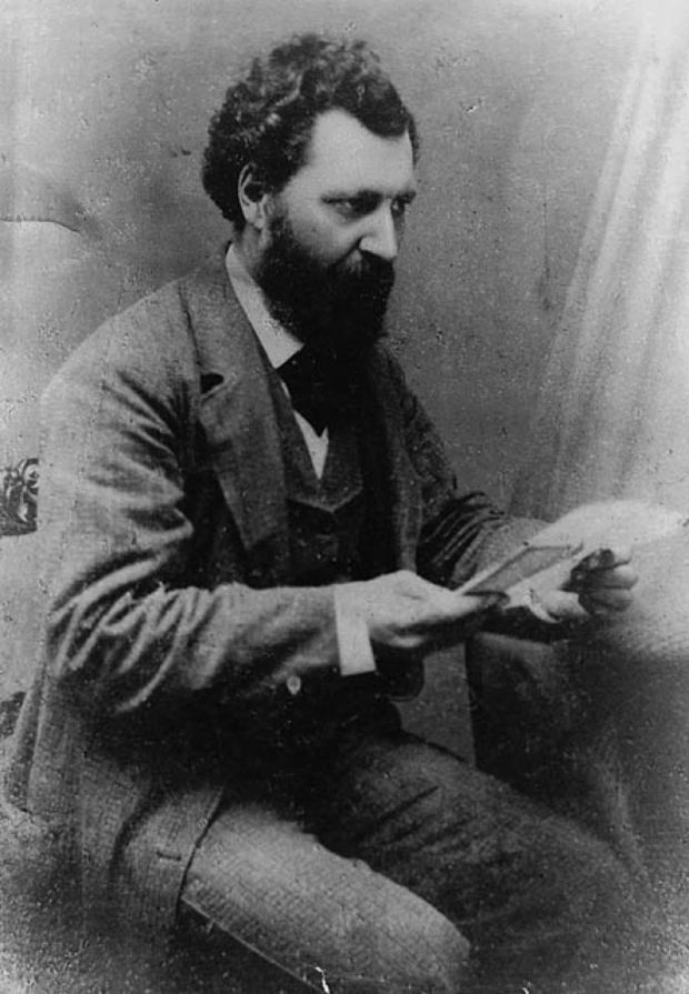 A black and white photograph of Louis Riel, seated, holding a piece of paper in his hand but not looking at it. He is bearded, has dark curly hair, and wears a three-piece woolen suit with a tie.