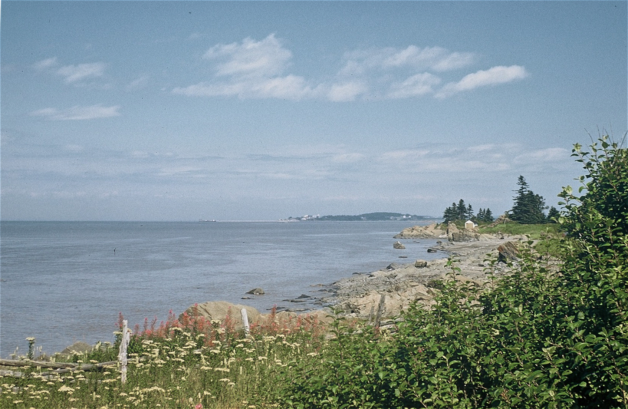 Colour photograph of a riverside scene at Rivière-du-Loup with a wooden fence in the foreground, rocks, sand, and shrubs as well as the river and some clouds in the sky.