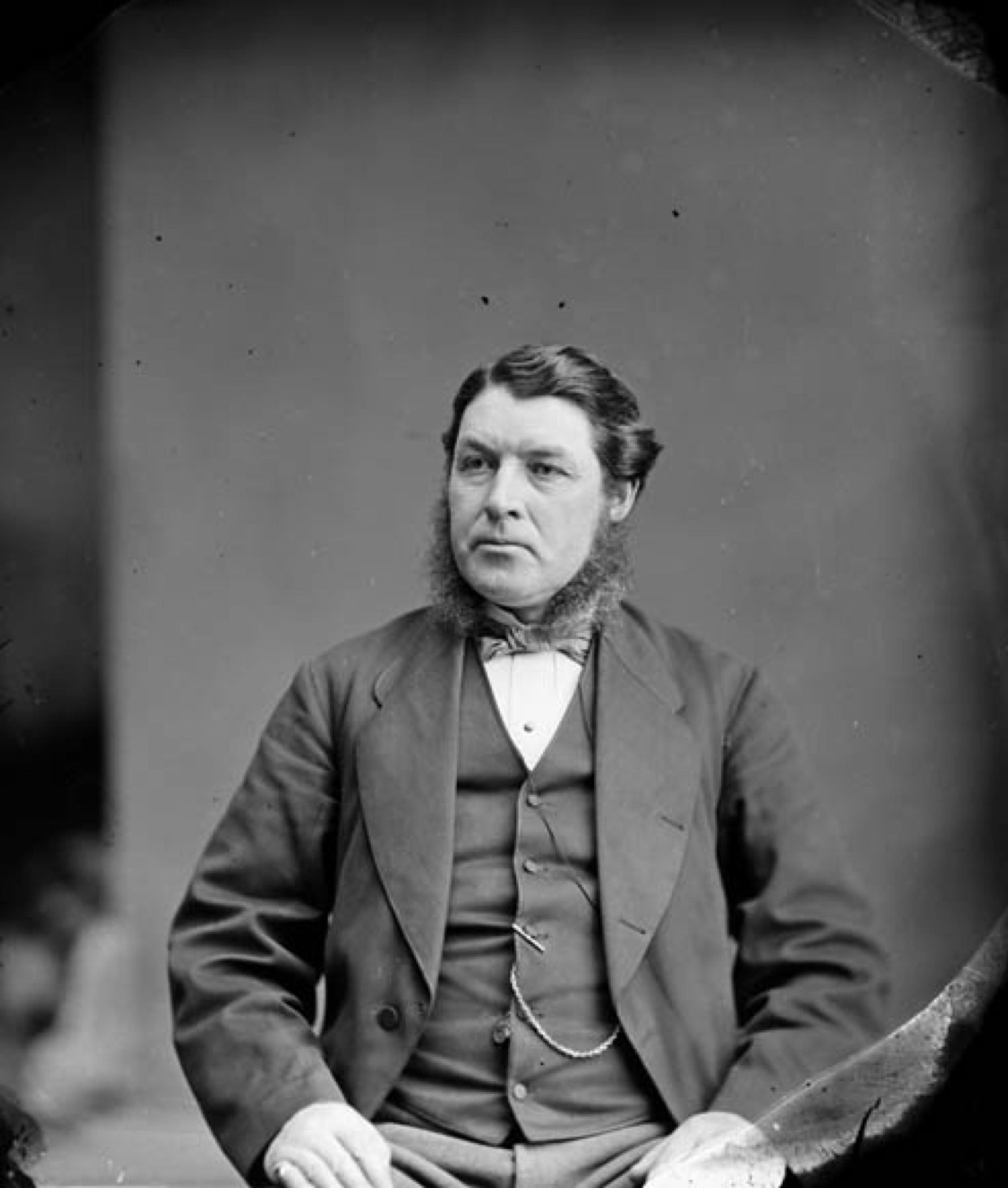 A formal black and white photograph of Sir Charles Tupper, seated, with dark hair and sideburns, in a suite and vest, his hands resting on his thighs and a watch-chain visible across his front. His expression is frowning and the edges of his mouth are turned down.