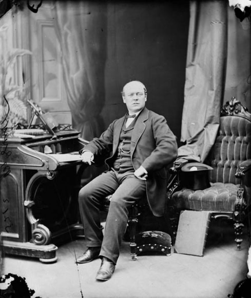 A black and white photo of Sir Thomas McGreevy, a middle-aged partially bald man sitting in an upholstered chair with one arm resting on the desk; he is facing the camera. The style of the dark formal clothing looks mid-Victorian; a top hat rests upside down on another upholstered chair next to the sitter. Draperies hang in the background, where there is a painted backdrop of a door and more drapery.
