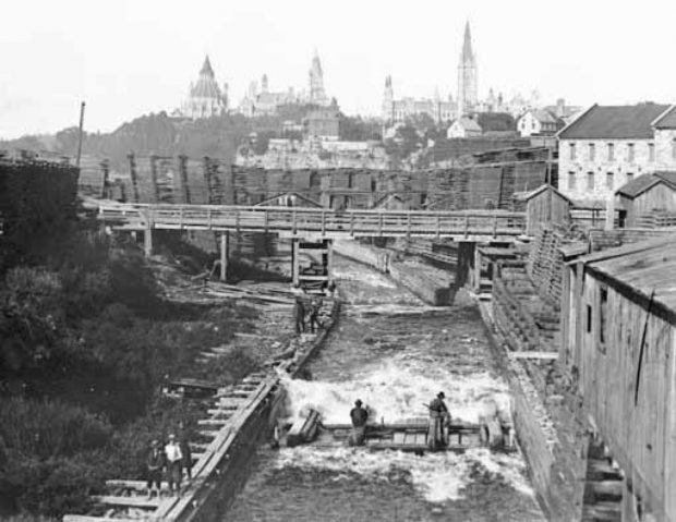Black and white photograph taken from above a canal with Ottawa's parliament buildings in the distance; in the canal are men on a logging raft.