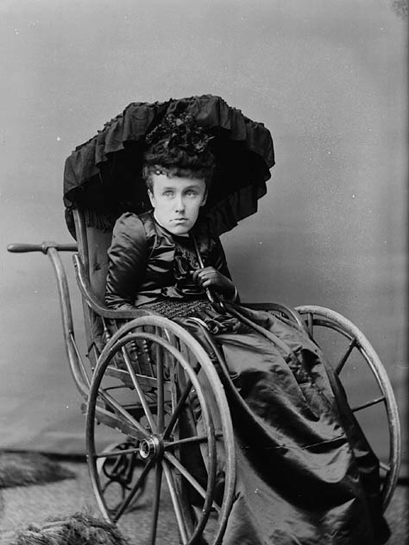 Black and white photo of an adult woman (Mary Macdonald) wearing a flat, vacant expression, sitting in a 19th century style wheelchair and holding a black parasol over her head.