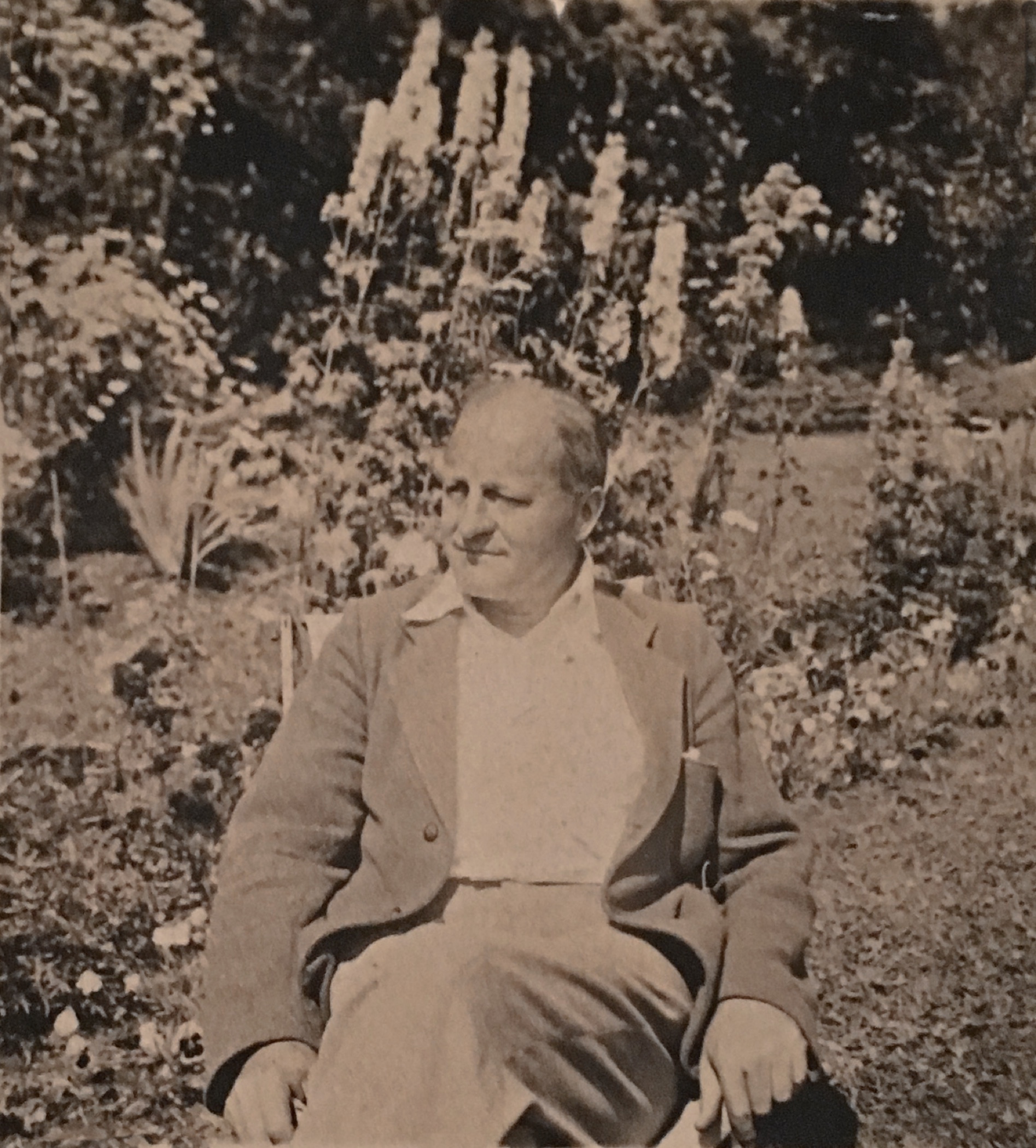 Sepia-toned photo of a middle-aged gentleman, Sir Herbert Symington, sitting on a chair outdoors with garden flowers (delphiniums) behind him.