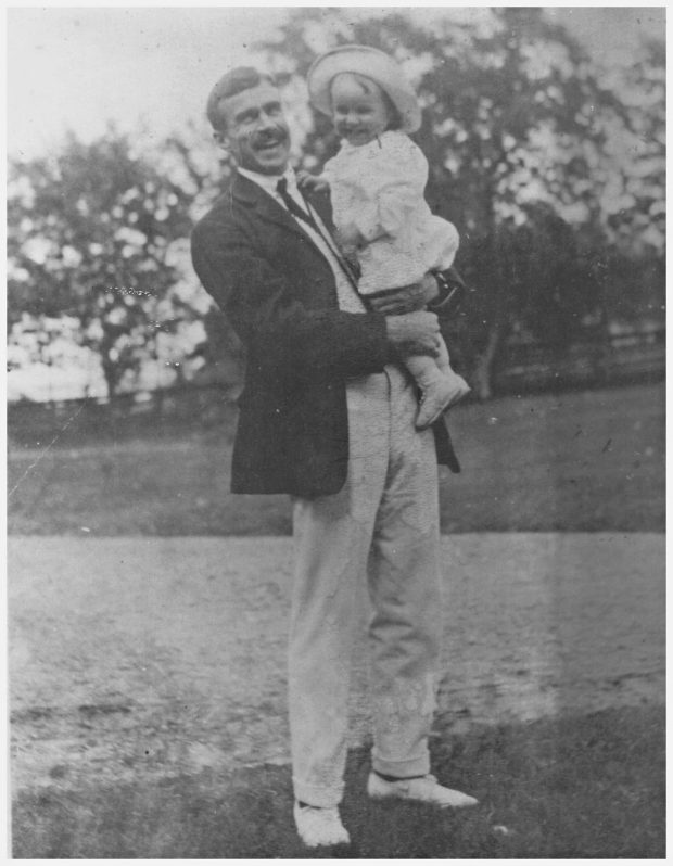 Black and white photograph of Kenneth Molson, a tall man smiling and holding a toddler, also smiling, posing outdoors with lawn and trees in the background.