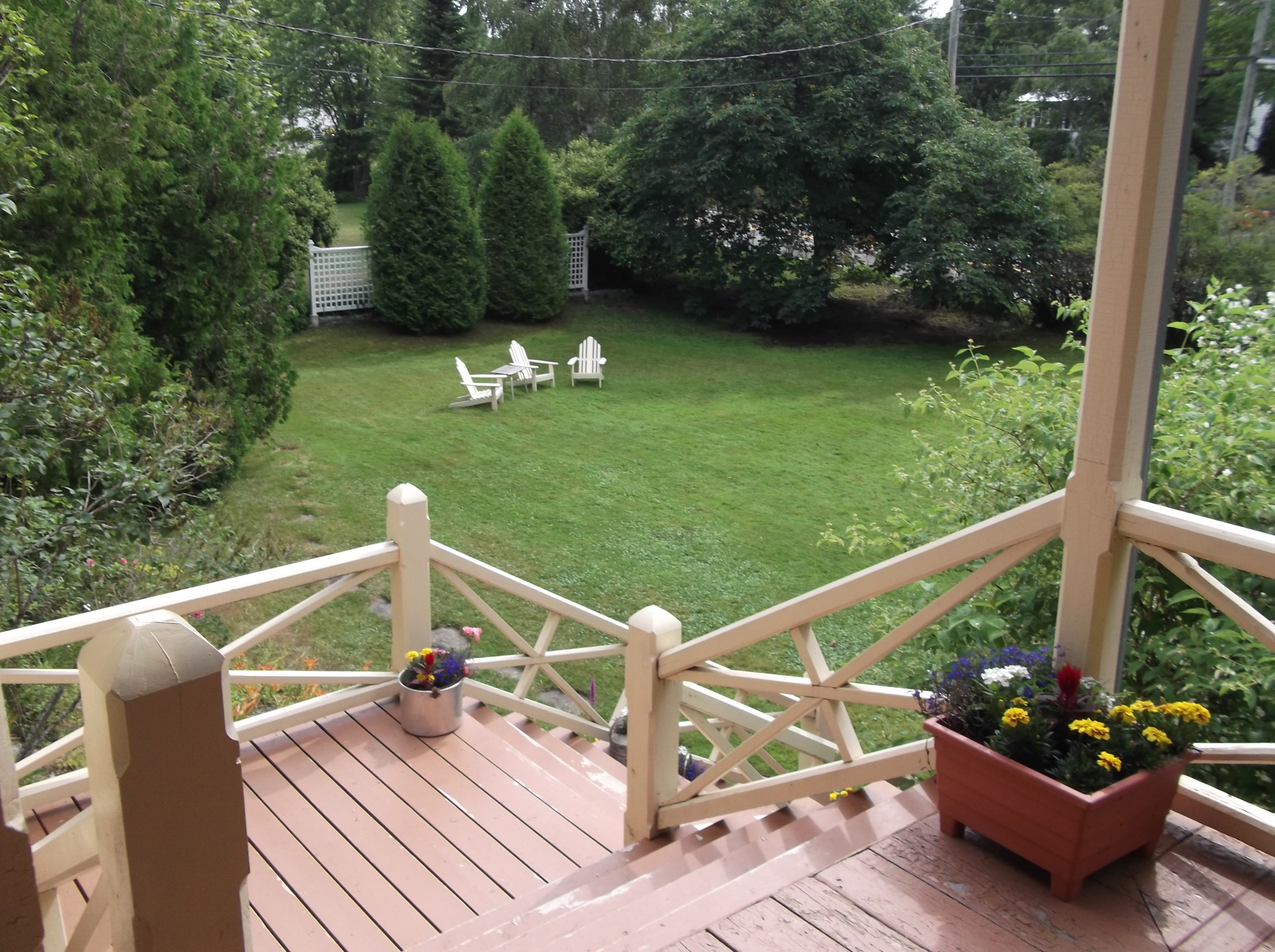 Colour photograph of the view of a well-maintained lawn in front of a large verandah, visible in the foreground. In the distance, on the lawn, are three empty white lawn chairs. The lawn is rimmed with cedar and fir trees.