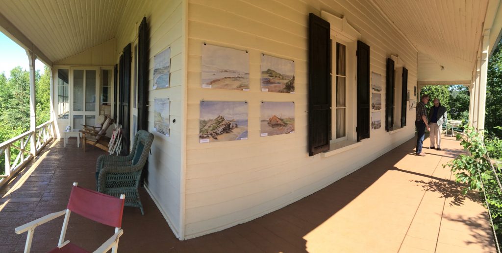 A colour photo depicting a wide view of two aspects of a large verandah, the shaded side with chairs on it and the sunny side with two people standing at the far end.