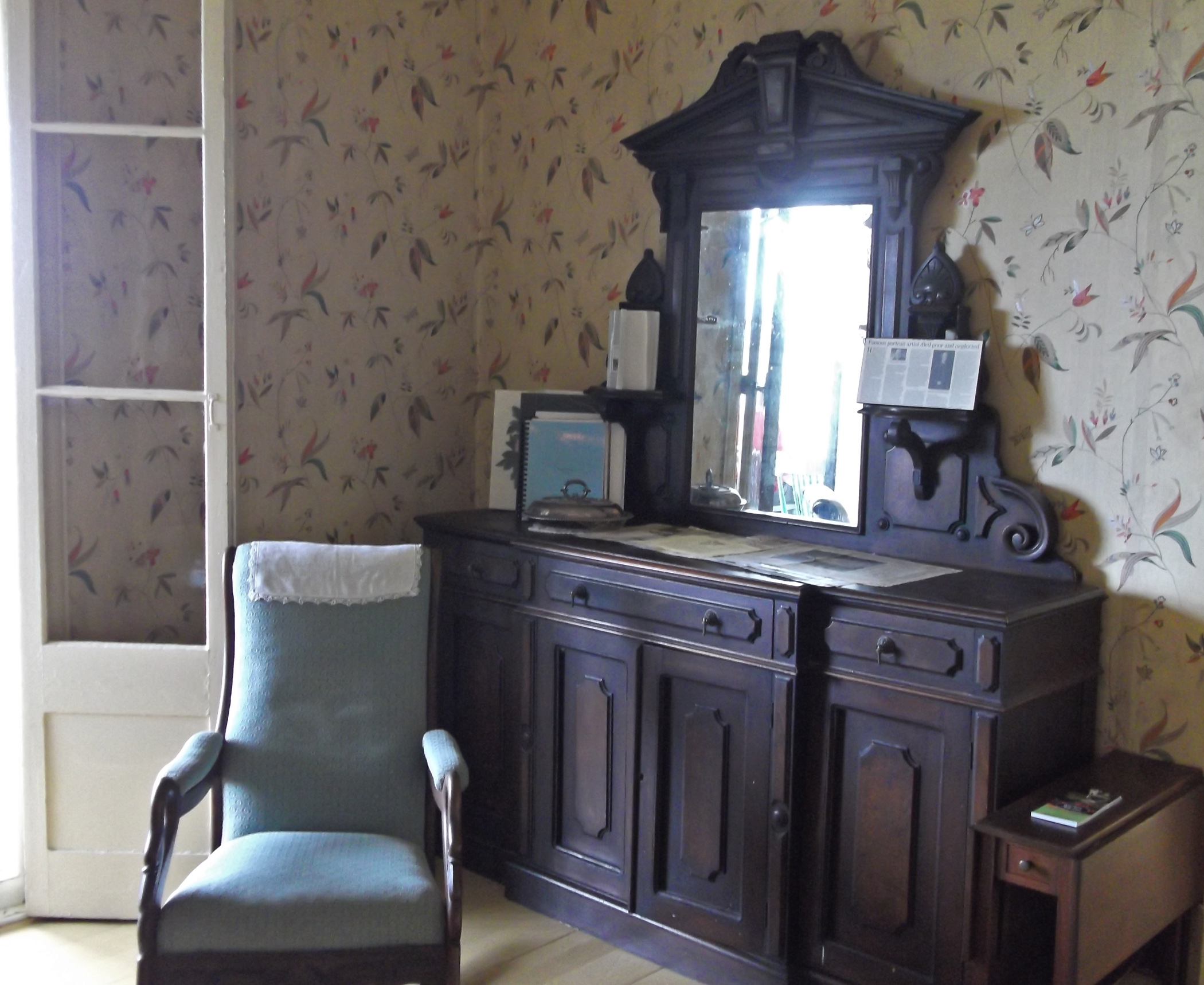 View of the interior corner of a dining room, with an upholstered chair and a wooden sideboard with drawers, cupboard doors and a mirror. The wall behind the furniture is wallpapered in a floral design.