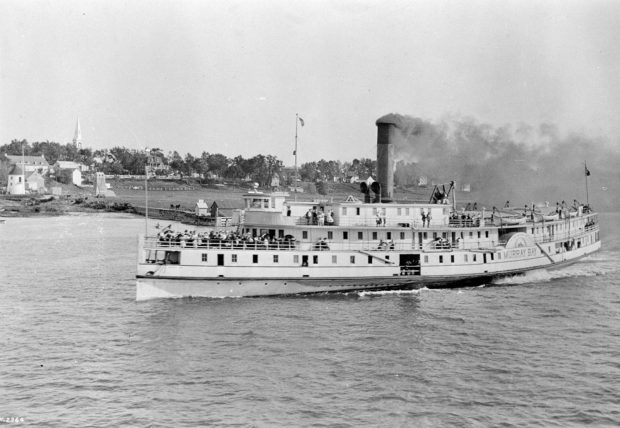 Black and white image of a medium-sized steamship ferry, in transit on a large river, with black smoke coming out of the smoke stack and the front deck crowded with people.