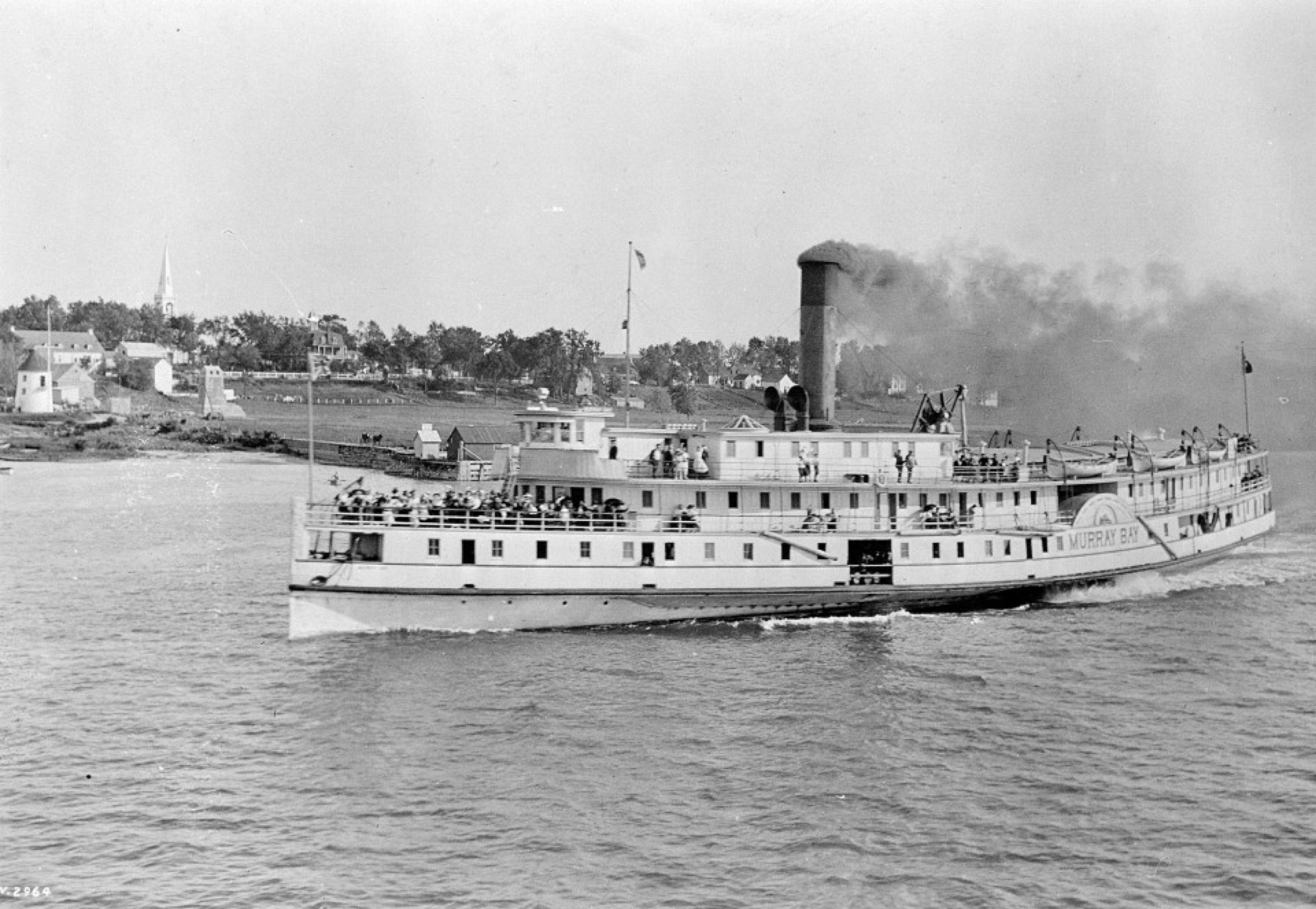 Black and white image of a medium-sized steamship ferry, in transit on a large river, with black smoke coming out of the smoke stack and the front deck crowded with people.