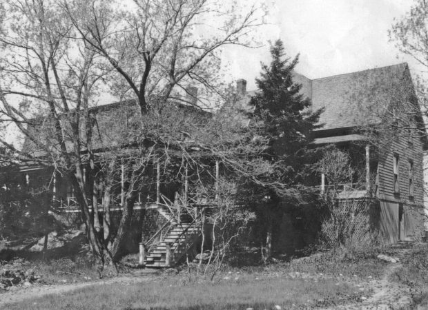 Black and white photograph of a large house (Villa Les Rochers) with a verandah spanning the front of it, a set of stairs leading up to the verandah, an overgrown deciduous tree and an evergreen tree in front.
