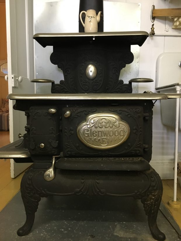 A colour photograph of a Glenwood brand cast iron cookstove, with polished metal cooking and warming surfaces. The viewer is looking at the stove from the front, facing the oven compartment's door. The stove sits on a square of tin that protects the floor. A ceramic coffeepot stands on the uppermost warming shelf of the stove.