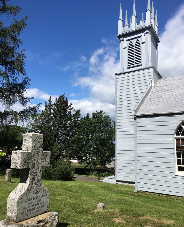 A colour, side-view photograph of St. Bartholomew's, an old wooden church with a front steeple. In the photo's foreground is a gravestone and in the background, trees and sky.