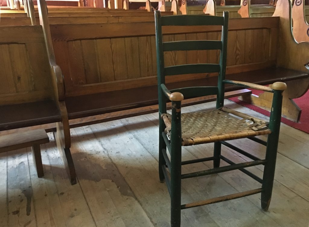 Photographie couleur d'une chaise en bois avec un siège à lattes, peinte en vert, dont on peut constater la vétusté par les lattes détachées du cadre sur un côté du siège ainsi que la peinture écaillée sur les appuie-mains des accoudoirs. La chaise se trouve devant une rangée de bancs d’église en bois. On aperçoit une surface du vieux plancher de bois franc brut. 