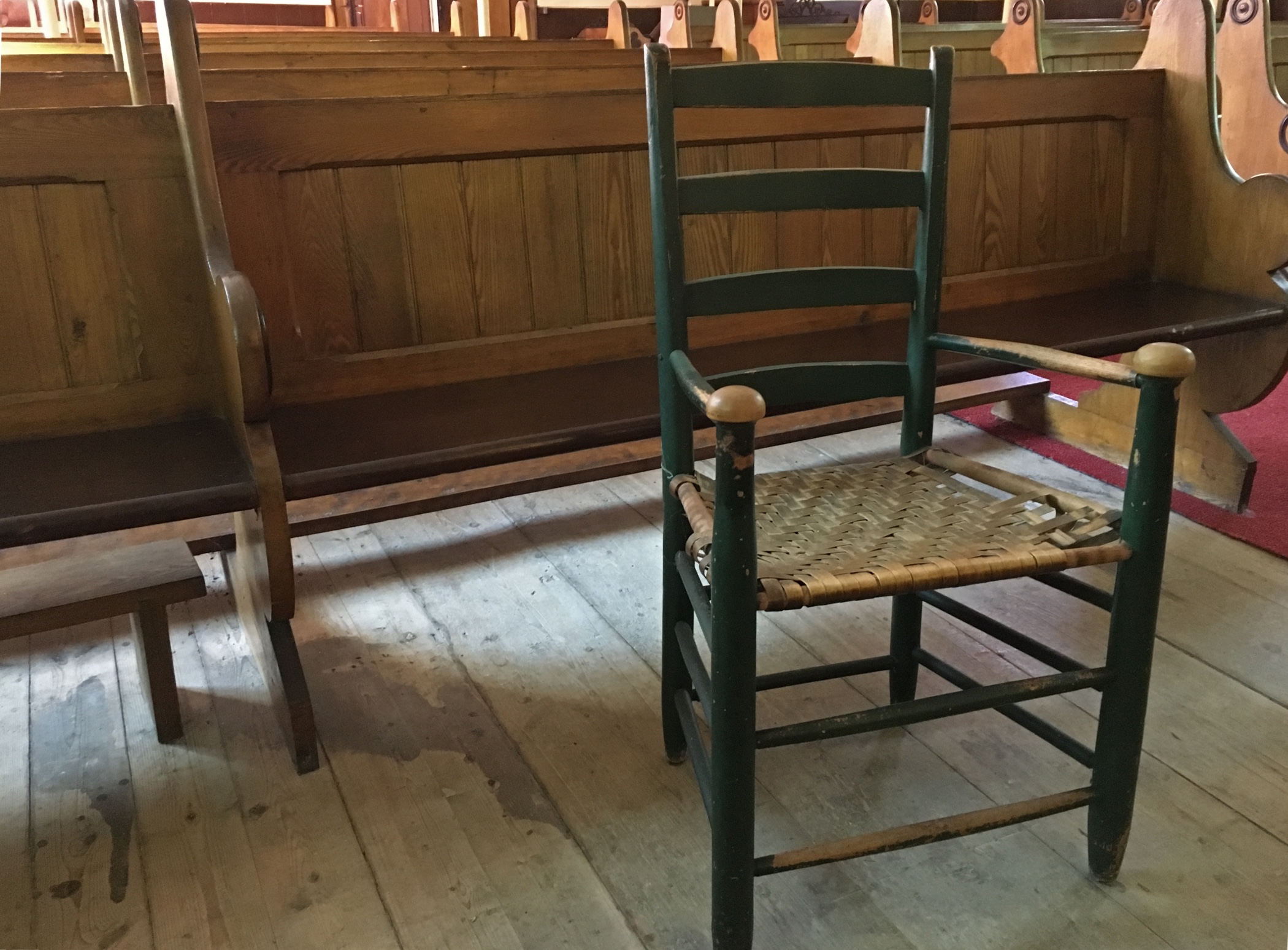 A colour photograph of a wooden slat-backed, painted chair with a woven seat which is so worn it is almost unattached to the frame of the seat on one side. The round-knob handles of the chair, and the armrests themselves, are so worn that all the paint has come off that part. The chair has been placed in front of a wooden pew behind which there are rows of pews. The wide floorboards of the church floor are unpainted and dry.