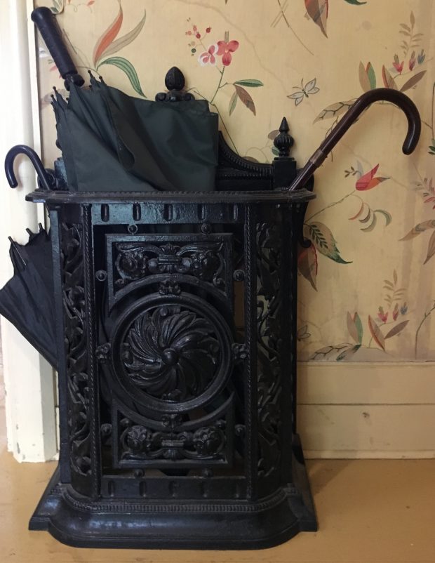 A cast-iron black painted umbrella stand about two and a half feet (62 cm) high and 18 inches (45 cm) wide, six inches (15 cm) deep. The cast iron is formed with symmetrical floral designs. The handles of two canes and one umbrella protrude from the top of the stand. Wallpaper adorns the wall behind it.
