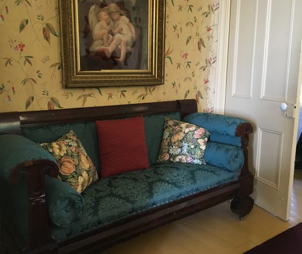 A colour photo of an old-fashioned upholstered chesterfield set in a nook behind a door, its back against a wallpapered wall upon which hangs a painting of cherubs.